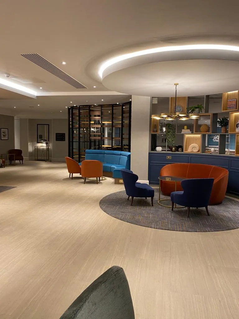 Open hotel lounge under a lit ceiling ring, with display shelving and armchairs