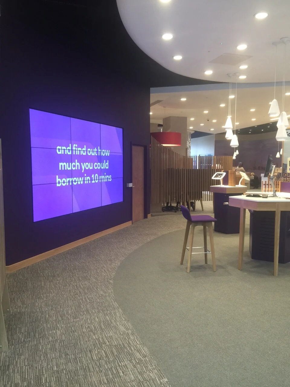 Purple video wall and oak-legged service desks under white pendant lights in the NatWest Crossrail Canary Wharf branch