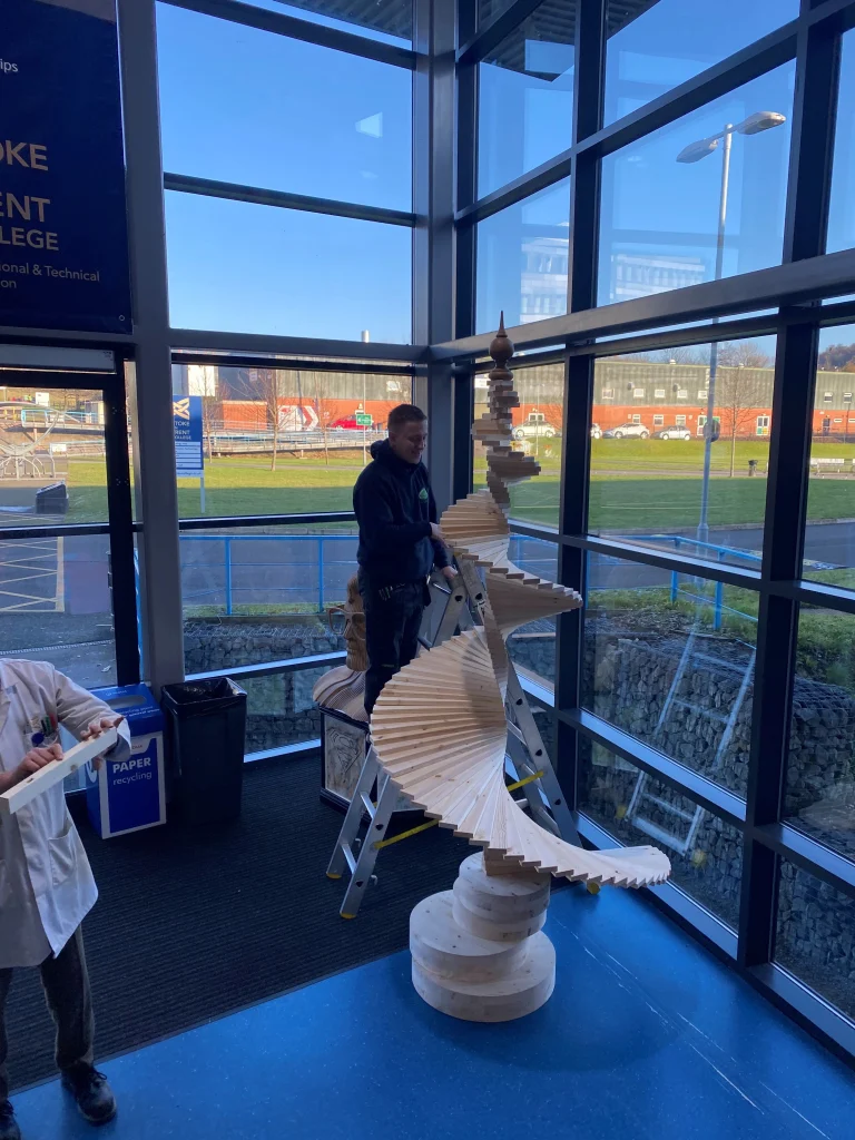 Carve Interiors apprentice assembling a tall spiral timber sculpture in the Stoke on Trent College foyer